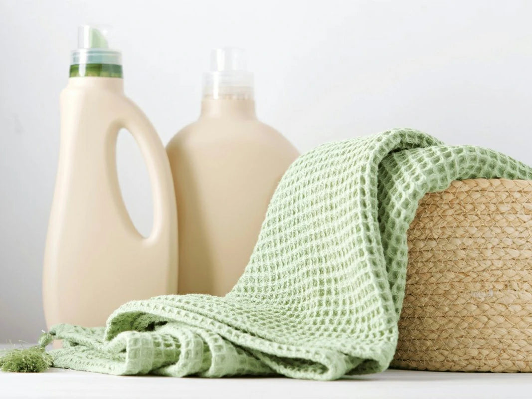 Two bottles of detergent with a green towel draped over a woven basket on a light background