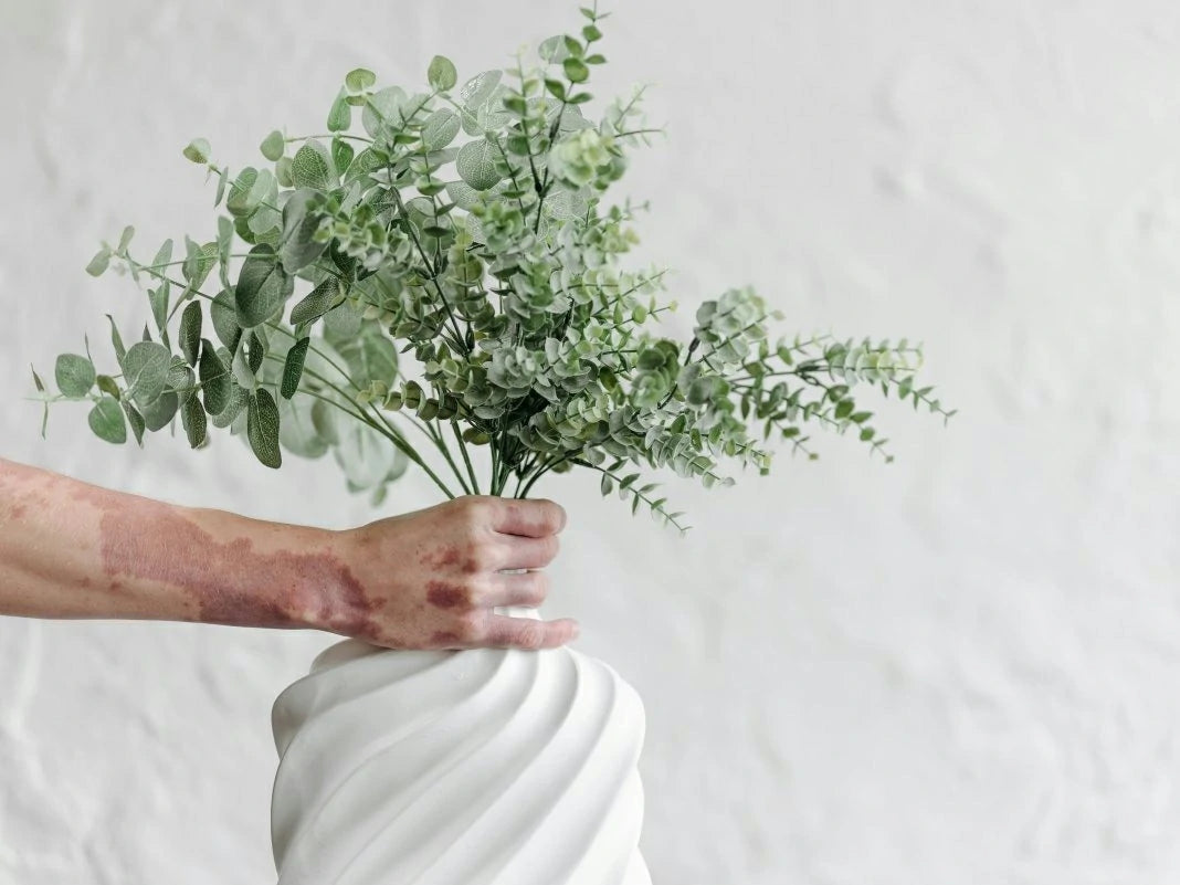 Hand holding a bouquet of greenery in a white vase against a light gray background